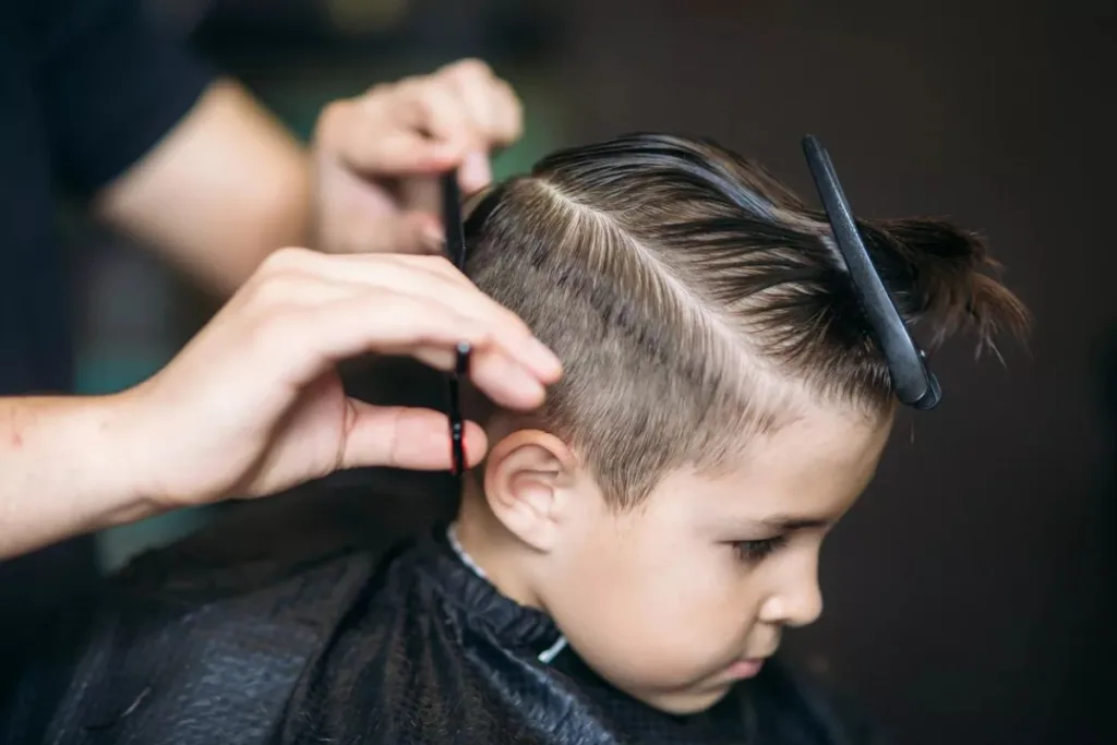 Um menino fazendo um corte de cabelo masculino infantil, com close das mãos de um barbeiro usando um pente no cabelo aparado da criança, com foco na lateral do cabelo